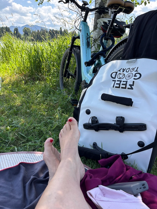 Eine Person macht eine Pause auf einer Decke im Grünen. Im Vordergrund sind die ausgestreckten Füße zu sehen, daneben zwei Fahrräder und eine weiße Fahrradtasche. Im Hintergrund liegen Wiese, Berge und ein blauer Himmel mit Wolken.nd einer Rad